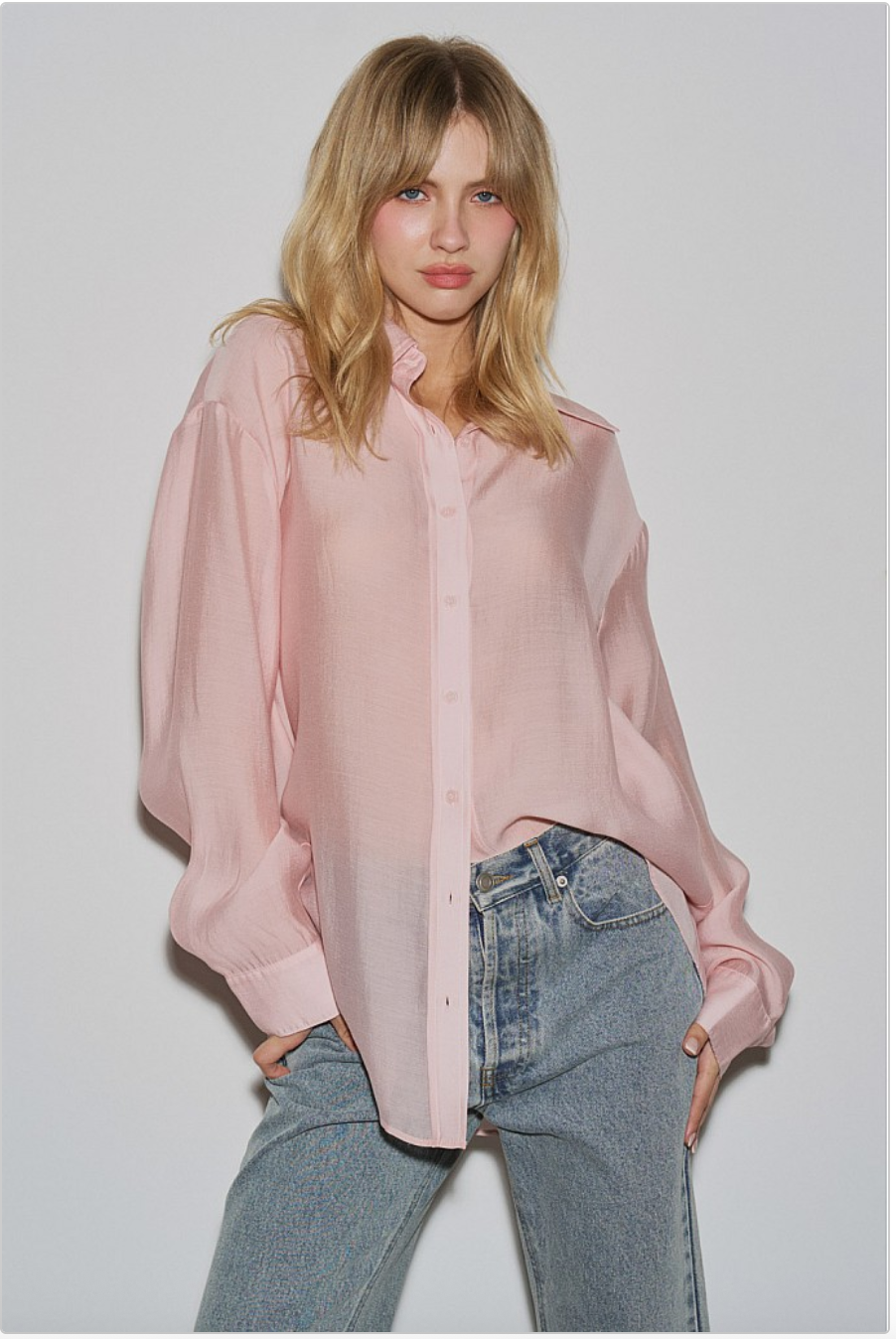 Model wearing a blush pink oversized sheer button-down shirt with light-wash jeans, front view against a white wall.