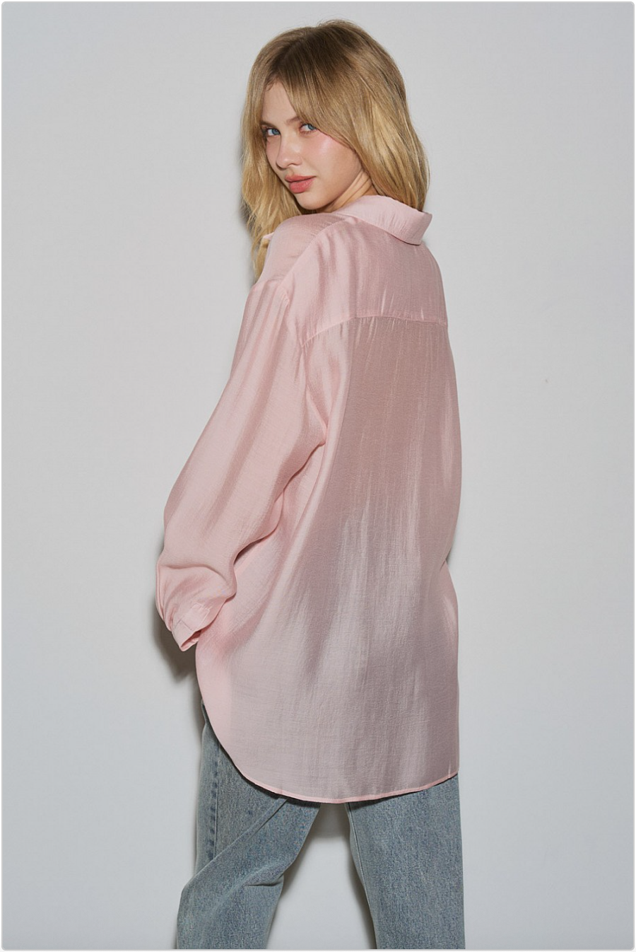 Back view of blush pink oversized sheer button-down shirt showing relaxed drape and longline hem, styled with jeans.